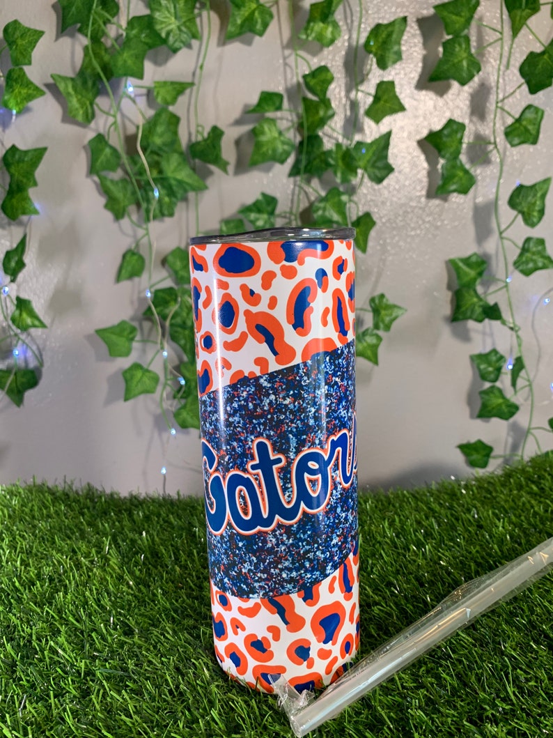 May include: A white, blue, and orange stainless steel tumbler with a leopard print design and the word "Gator" in blue lettering.