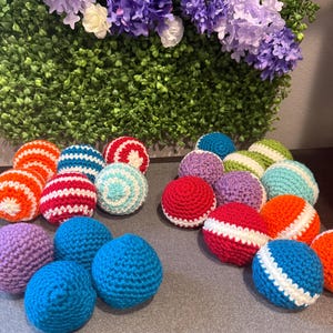 May include: A collection of small, round, crocheted yarn balls in various colors including red, orange, blue, purple, and teal. Some balls feature solid colors, while others display horizontal or spiral striped patterns in contrasting white yarn.