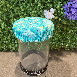 May include: A clear glass jar with a blue floral fabric lid. The lid has a pattern of small blue and yellow flowers on a turquoise background. A black band with white text is wrapped around the jar.