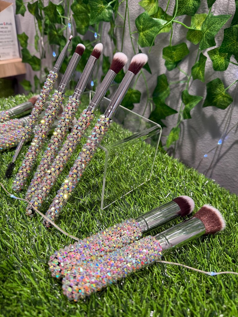May include: A collection of makeup brushes with silver metal handles and sparkling, iridescent rhinestone-covered grips. The brush heads are a mix of white and dark brown. The brushes are arranged on a clear acrylic stand and green grass.