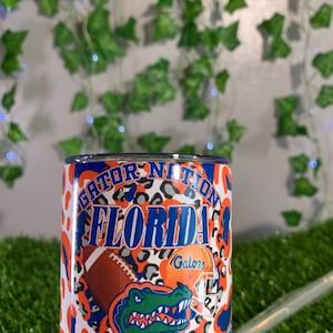 May include: A white tumbler with an orange and black leopard print design. The tumbler features the words "Gator Nation Florida Gators" and a green alligator with a blue and white helmet.