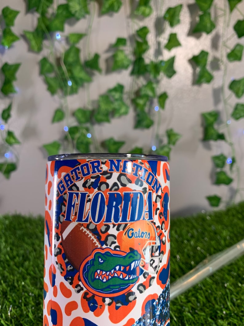 May include: A white tumbler with an orange and black leopard print design. The tumbler features the words "Gator Nation Florida Gators" and a green alligator with a blue and white helmet.