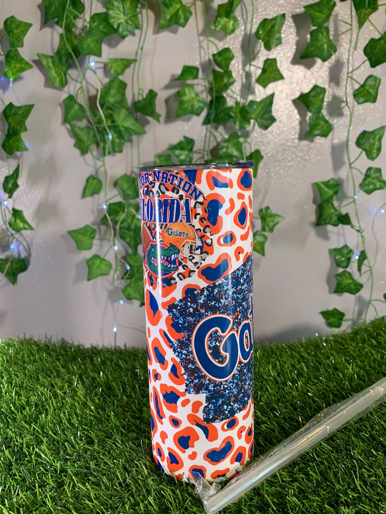 May include: A stainless steel tumbler with a white, orange, and blue leopard print design. The tumbler features the text "Gator Nation Florida" and a gator logo. The tumbler also has a blue and white glitter design with the text "Gators".