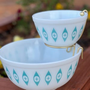May include: A white glass serving bowl set with a teal and white geometric pattern. The set includes a large bowl and a smaller bowl that sits on a gold metal stand.