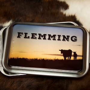 May include: A silver metal baking pan with a sunset scene and the text "FLEMMING" printed on it. The scene features two black silhouettes of cows in a field.