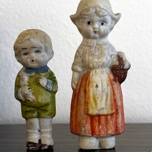 May include: Two porcelain figurines, a girl wearing a red and white dress and a boy wearing a green and blue outfit. The girl is holding a basket and the boy is holding a white bird.