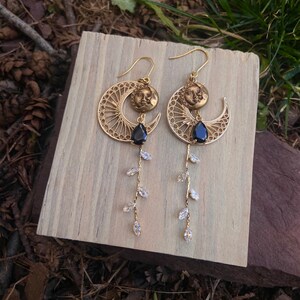 May include: Gold earrings with a crescent moon design featuring a sun and moon face charm. The earrings have a dangling chain of clear crystal teardrop-shaped beads.