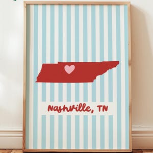 May include: A framed digital print featuring a light blue and white vertical striped background. Centered is a red silhouette of the state of Tennessee with a small pink heart over Nashville. The text Nashville, TN is printed in red below.
