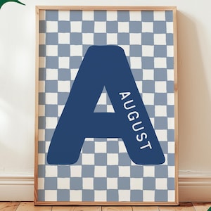 Op de afbeelding: Een rechthoekige wandkunstprint met een grote, donkerblauwe letter A gecentreerd op een lichtblauwe en witte geblokte achtergrond. Het woord AUGUST is in witte hoofdletters verticaal langs de rechterkant van de letter A gedrukt.
