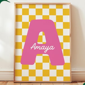 Op de afbeelding: Een ingelijste print met een roze letter 'A' en de naam 'Amaya' in witte letters, tegen een geel en wit geblokte achtergrond. De lijst is licht van kleur. Een groene tekstballon zegt 'Custom Digital Download'.
