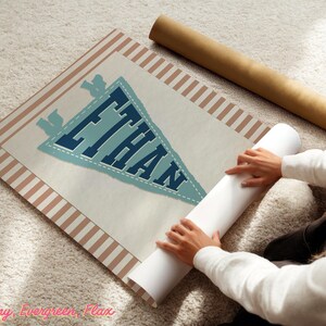 May include: A personalised pennant-style print with the name "ETHAN" in blue lettering. The print has a light blue pennant shape with a white border and is set against a cream background with a striped border. A rolled-up poster is next to the print.