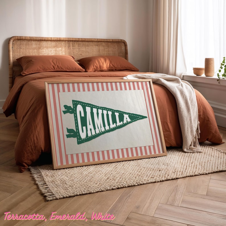 May include: A framed pennant with the name "CAMILLA" in white lettering on a green triangle. The pennant is set against a pink and white striped background. The frame is light brown. The bed has terracotta bedding.