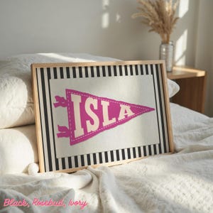 May include: A framed print featuring a pennant design with the name "ISLA" in rosebud pink. The pennant is set against a cream background with a black and white striped border. The frame is made of light-coloured wood.