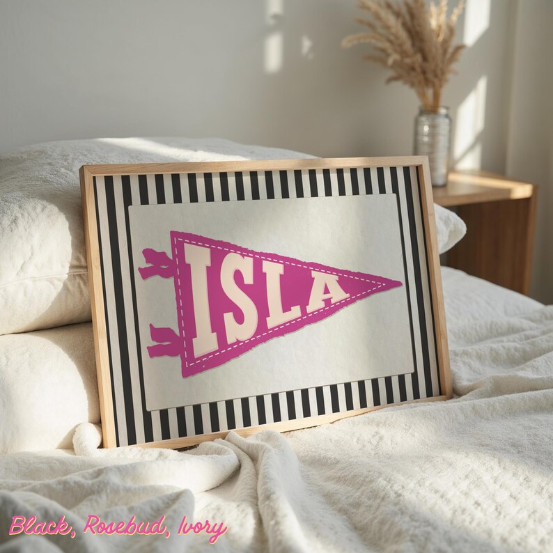 May include: A framed print featuring a pennant design with the name "ISLA" in rosebud pink. The pennant is set against a cream background with a black and white striped border. The frame is made of light-coloured wood.