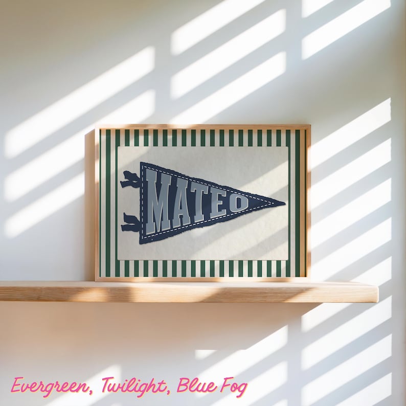May include: A framed pennant with the name "MATEO" in grey lettering on a navy blue pennant. The pennant is framed with a green and white striped border and a wooden frame. The pennant is displayed on a wooden shelf.