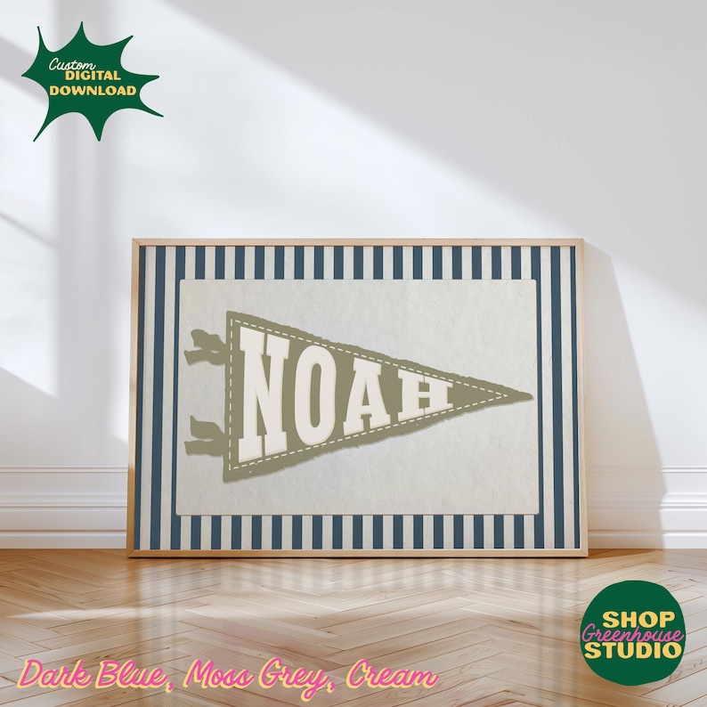 May include: A framed print with a pennant design displaying the name "NOAH". The pennant is moss grey with a cream background and a dark blue striped border. The frame is light wood. The print is suitable for a child's room.
