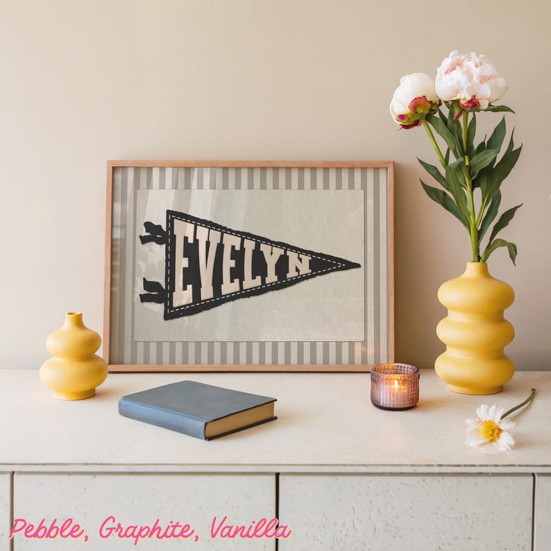 May include: A framed pennant with the name "EVELYN" in white letters on a black and white pennant. The pennant is displayed on a white surface with a book, two yellow vases, a candle, and flowers.