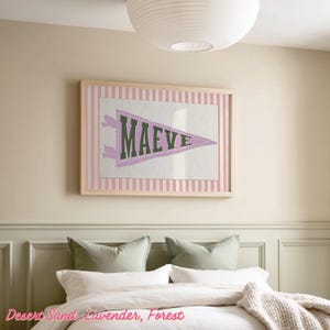 May include: A framed pennant-style wall art piece with the name "MAEVE" in dark green letters on a lavender pennant. The pennant is set against a white background with a pink striped border, all within a light wood frame. The artwork hangs above a bed.