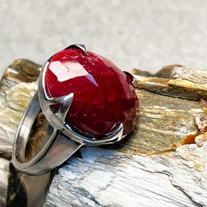 May include: A silver ring featuring a large, faceted, oval-shaped ruby gemstone. The deep red ruby is set in a prong setting, and the ring band is a simple, polished silver. The ring is displayed on a textured, natural stone surface.