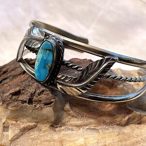 May include: A silver cuff bracelet featuring a central oval turquoise stone with dark matrix veins. The band is accented with two detailed silver feathers on either side of the stone and a twisted silver wire detail along the open cuff design.