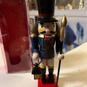 Peut inclure: Une figurine de casse-noisette en bois avec un chapeau noir, un uniforme bleu et une lance dorée. Le casse-noisette tient une lanterne dans une main et une lance dans l'autre. Le casse-noisette est debout sur une base rouge.