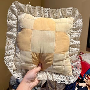 May include: Square patchwork pillow with a cream-colored lace trim. The pillow features a central design of light peach and cream-colored squares. The lace trim adds a vintage, handcrafted aesthetic to the pillow.