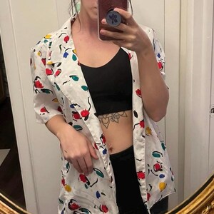 May include: A white button-up shirt with a colorful abstract print. The shirt has a relaxed fit and is open at the front, revealing a black tank top underneath.
