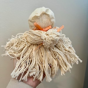 May include: A handmade doll made from white string with a white fabric bonnet and a peach-colored ribbon tied around the head.