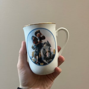 Puede incluir: Taza de cerámica blanca con borde dorado y una ilustración azul y blanca de una niña montando a caballo. La ilustración está rodeada de un círculo azul.
