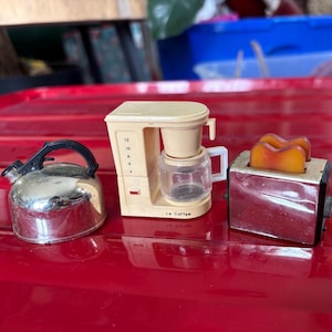 May include: A miniature kitchen set featuring a silver kettle, a beige coffee maker with a clear carafe, and a red toaster with two slices of toast. The coffee maker has the text "Le Coffee" on the front.