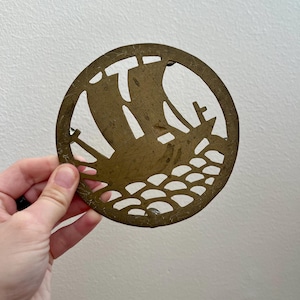 May include: A round brass coaster with a cutout design of a ship sailing on the water. The coaster is about 4 inches in diameter.