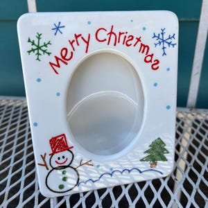 May include: White ceramic Christmas picture frame with an oval aperture. The frame is decorated with a snowman, snowflakes, a Christmas tree, and the words "Merry Christmas" in red. The frame is on a white metal surface.