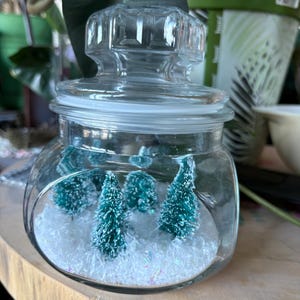 May include: A clear glass jar with a lid, filled with white faux snow and small, green Christmas trees. The jar is sitting on a wooden surface, with green foliage in the background.