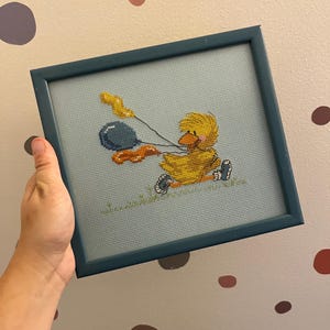 K&ouml;nnte beinhalten: Ein gerahmtes Kreuzstichbild mit einem gelben Entenk&uuml;ken, das blaue und orangefarbene Ballons h&auml;lt. Das K&uuml;ken tr&auml;gt eine rote Schleife und sitzt auf einem gr&uuml;nen Grasfleck.