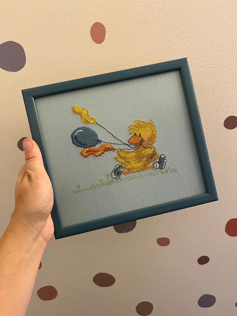 K&ouml;nnte beinhalten: Ein gerahmtes Kreuzstichbild mit einem gelben Entenk&uuml;ken, das blaue und orangefarbene Ballons h&auml;lt. Das K&uuml;ken tr&auml;gt eine rote Schleife und sitzt auf einem gr&uuml;nen Grasfleck.