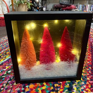 May include: A black framed shadow box featuring three bottle brush Christmas trees in shades of orange and red, set against a backdrop of warm fairy lights and faux snow. The box sits on a colorful braided rug.