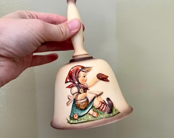 May include: Cream-colored ceramic bell with a painted illustration of a young person wearing a red headscarf and a green apron. The bell has a light brown handle and a detailed design of a basket and flowers.