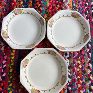 May include: Three white octagonal plates with floral designs in pink, yellow, and green. The plates have a decorative border and are set against a colorful, woven background. These plates are suitable for serving food.