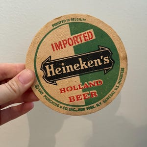 May include: A round, brown paper beer coaster with green and black text that reads "IMPORTED Heineken's HOLLAND BEER" and "Printed in Belgium". The text is surrounded by a white border.