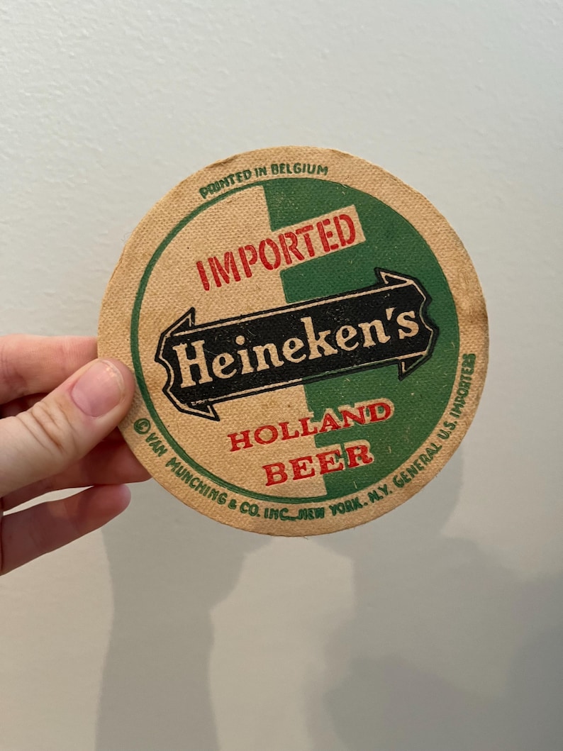 May include: A round, brown paper beer coaster with green and black text that reads "IMPORTED Heineken's HOLLAND BEER" and "Printed in Belgium". The text is surrounded by a white border.