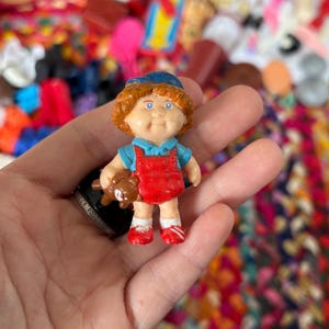 May include: A small, vintage Cabbage Patch Kids figurine. The doll has orange hair, blue eyes, and is wearing a blue shirt, red overalls, and a blue hat. It is holding a small brown teddy bear.