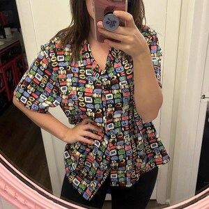 May include: A short-sleeved button-up shirt with a colorful geometric pattern. The shirt has a V-neck and a black background with multi-colored squares and rectangles.