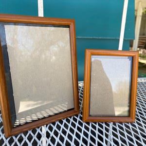 May include: Two wooden picture frames, one rectangular and one smaller, are displayed on a white metal surface. The frames have a warm, brown finish and are empty, with a neutral-colored backing.