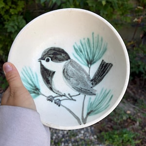 Puede incluir: Un cuenco de cerámica blanco con un diseño de pájaro pintado a mano. El pájaro está representado en tonos grises y negros, posado en una rama con agujas de pino de color verde azulado. El cuenco está sostenido en una mano.