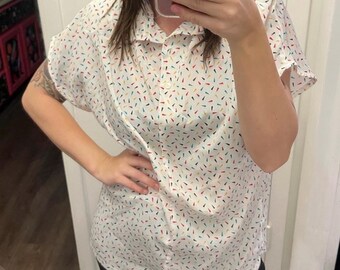 May include: White button-down shirt with a colorful confetti pattern. The short-sleeved shirt features a classic collar and a button-up front. The confetti design includes red, yellow, blue, and green accents, creating a playful and vibrant look.