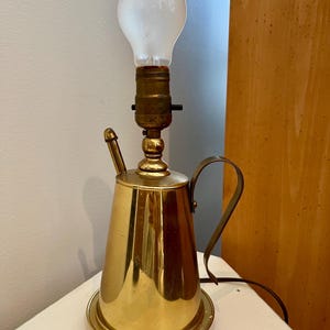 May include: A brass table lamp shaped like a teapot with a clear light bulb. The lamp has a black cord and a black switch.