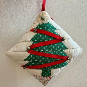 May include: A handmade quilted Christmas tree ornament with red ribbon and silver bells. The ornament is made of white and green fabric with a small white flower on top.