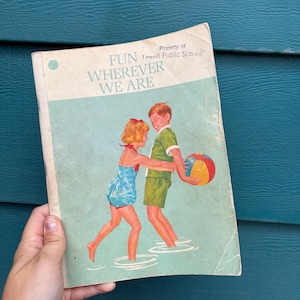 May include: Vintage book cover with the title "FUN WHEREVER WE ARE" and an illustration of two children playing with a beach ball. The cover is light blue and white with a retro design. The book is held by a hand.