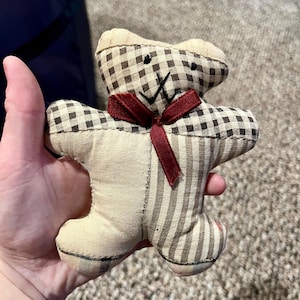 Op de afbeelding: Een patchwork teddybeer met een rode strik. De beer is gemaakt van bruin en wit geruit stof en gestreept stof.