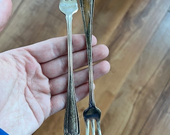May include: Two antique silver forks with ornate handles and tines. One fork has three tines, the other has four. The forks are held in a hand against a wooden background. The forks are likely for serving or eating.